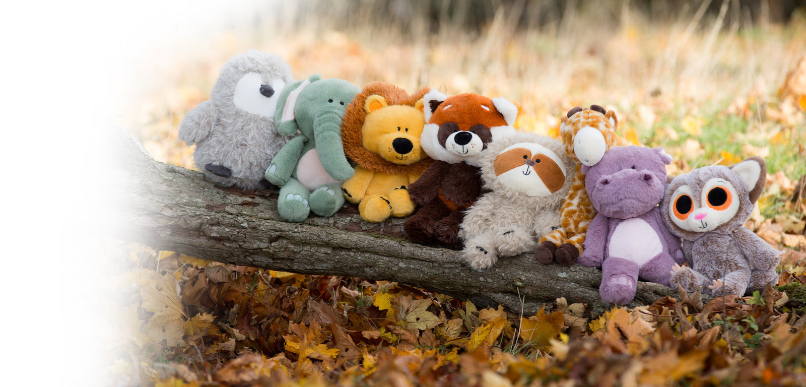 A range of MBI and ZSL plush toys sitting on a log. Penguin, elephant, lion, red panda, sloth, giraffe, hippo and bush baby.